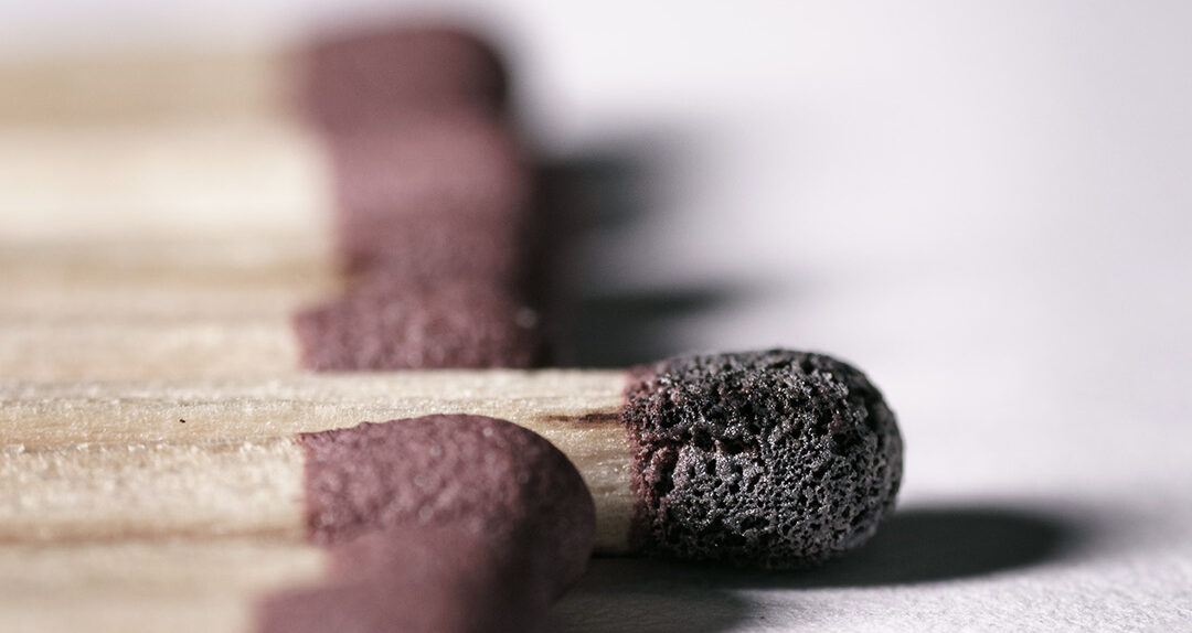 Streichhölzer von oben nach unten das Bild mittig teilend. Eins ragt mit abgebrannten Zündkopf aus der Reihe hervor.